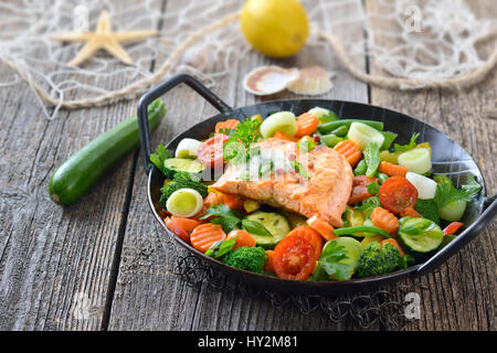 Fried salmon fillet on mixed colorful vegetables served in a frying pan,  a lemon and a fishing net with a starfish and seashells in the background Stock Photo