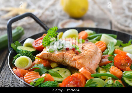 Fried salmon fillet on mixed colorful vegetables served in a frying pan,  a lemon and a fishing net with a starfish and seashells in the background Stock Photo