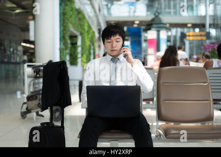 Asian businessman using smartphone and laptop while waiting for his flight in terminal at airport. Stock Photo