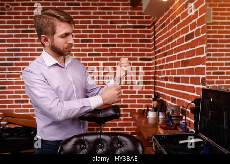 Portrait of the hairdresser. Cheerful young bearded man fastens the buttons on the sleeve on his shirt. Stock Photo
