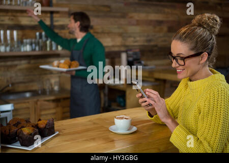 Happy woman using mobile phone while barista working in background at coffee shop Stock Photo