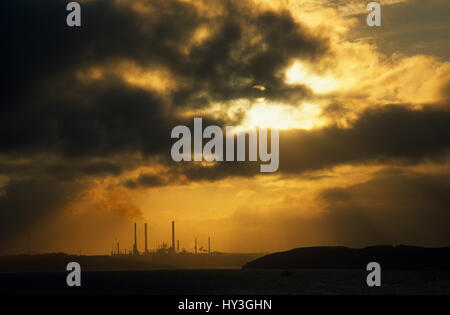 Wales, Pembrokeshire, Milford Haven, Distant view over sea towards Texaco oil refinery in golden light with low grey cloud. Stock Photo