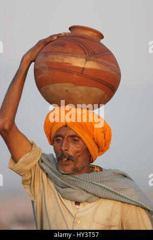 Indian man in orange turban carrying pot of water on his head at the Pushkar Fair in Rajasthan, India Stock Photo