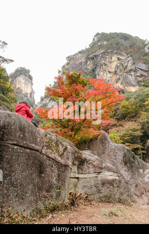 Woman in red jacket takes photo of autumn colour trees at bottom of rocky mountain in Shosenkyo Gorge, Japan Stock Photo