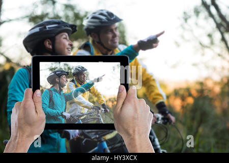 Cropped hand holding digital tablet against biker couple with mountain bike pointing in distance Stock Photo