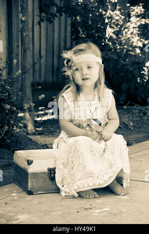 Sad little girl sitting on luggage, barefoot, wearing an old fashioned lacey dress. Stock Photo