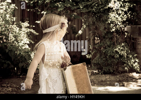 Little blonde toddler girl with peasant dress and luggage looking back over shoulder Stock Photo