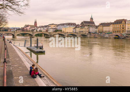 Teenager couple sitting down on Basel riverfront opposite Basel's Altstadt ('old town') on the footpath along the Rhine River. Stock Photo