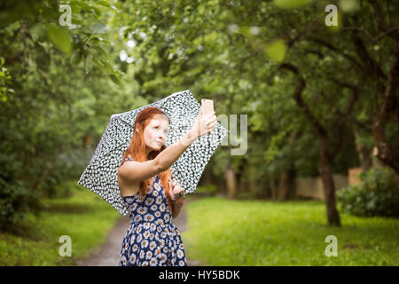 Finland, Pirkanmaa, Tampere, Woman wearing floral dress standing with umbrella in park and taking selfie