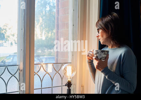 Sweden, Woman looking through window, holding coffee cup