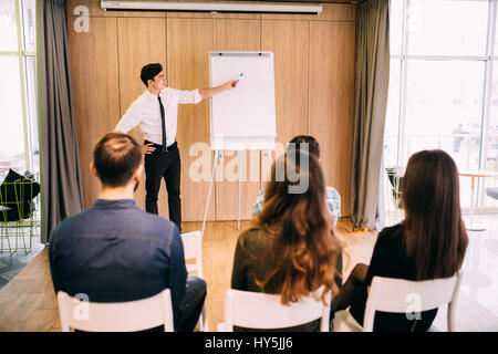 Rear View of Young Office Workers in Casual Outfits Listening to a Top Manager Explaining Something Using Illustrations in office Stock Photo
