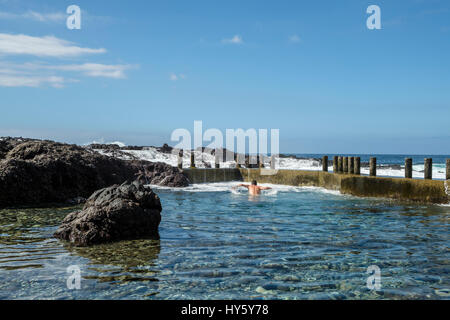 A man enjoys a refreshing swim in the still clear blue waters of a natural rock pool on the Tenerife coast near Alcala while waves from the Atlantic O Stock Photo