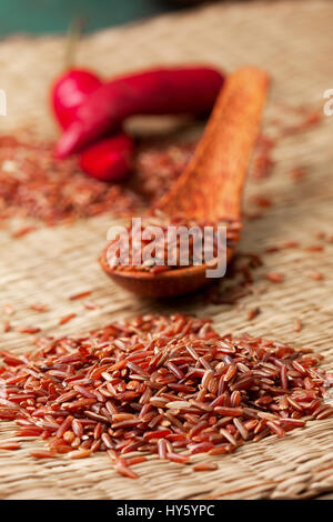 red rice in a wooden spoon, rice and red pepper on a straw background