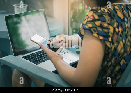 Asian woman using smartphone at home while working with laptop computer. Online business and wireless working from anywhere concept with vintage filte Stock Photo