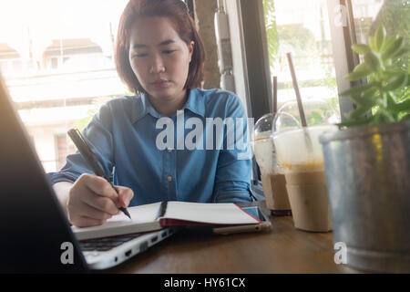 Young Asian hipster woman writing journal on notebook with serious emotion in coffee shop. Freelance business starup activity concept Stock Photo