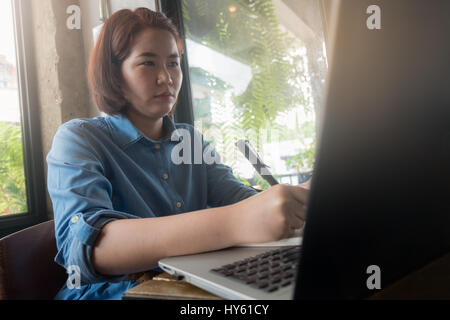 Young Asian hipster woman writing on notebook while working with laptop computer in coffee shop. Freelance business starup activity concept Stock Photo