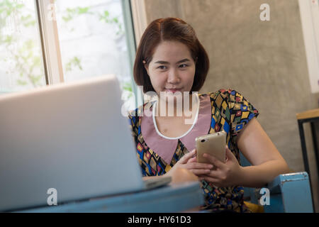 Asian woman using smartphone at home while working with laptop computer. Online business and wireless working from anywhere concept Stock Photo