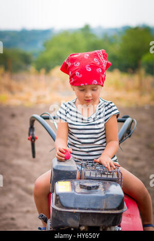 child in red bandana and stripe tee sits on tiller on the field. baby girl driver cultivator. Stock Photo