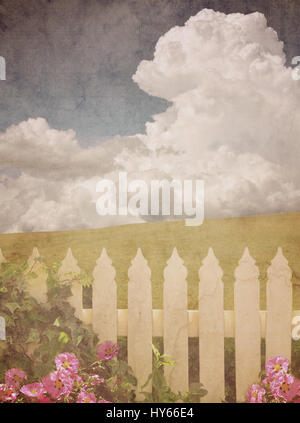 White picket fence with ivy and pink flowers in front of a green grassy hill and blue sky cloudscape background. Vintage paper textured image. Stock Photo