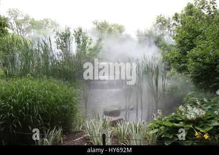 Fog hangs over a waterfall on a small river among stones and green bushes. Rocky bed of a small stream in impenetrable thickets. Smoke on the water. Stock Photo