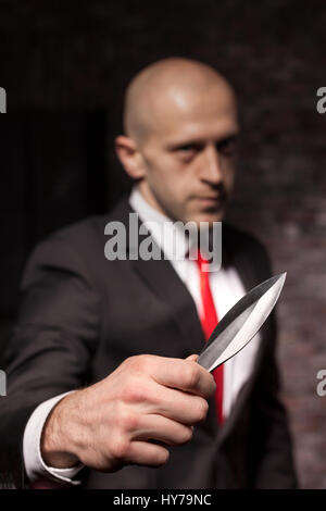 Silent killer, oriental martial arts in action. Bald contract murderer in suit and red tie holds combat knife. Fighting knife is a dangerous weapon in Stock Photo