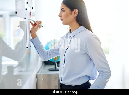 Side view on serious young adult woman in long sleeve blue blouse giggling while writing on white board with bright background Stock Photo