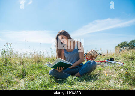 A young woman reads a book whilst sitting in a meadow Stock Photo