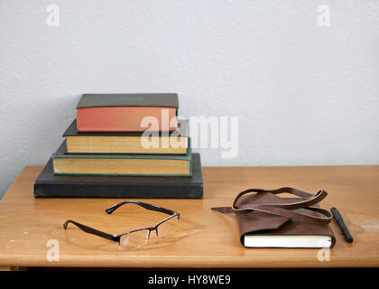 Leather journal, pencil, readers, and hardbound, used books stacked on a weathered, used wooden desk, against white wall - unplugged study concept Stock Photo