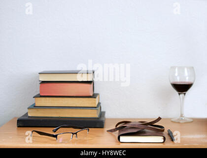 Leather journal, pencil, readers, and hardbound books stacked, used books on a weathered, used wooden desk, against white wall, and a glass of red win Stock Photo
