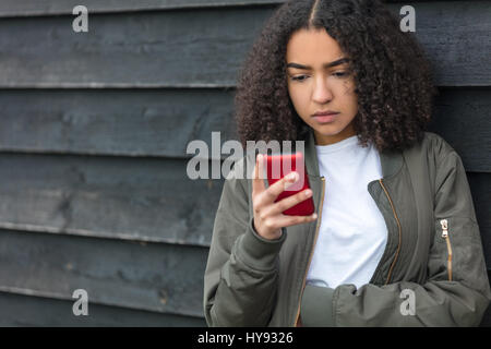 Portrait of beautiful sad depressed mixed race African American girl teenager female young woman texting on red cell phone wearing green bomber jacket Stock Photo