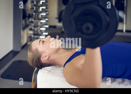 Close-up side view of young blonde woman in blue top doing bench pressing exercise with bar-bell in gym Stock Photo