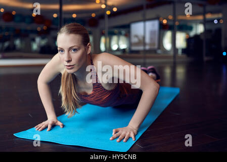 Young blonde woman doing push-up exercise in gym and looking at camera while lying on blue yoga carpet over mirror background Stock Photo