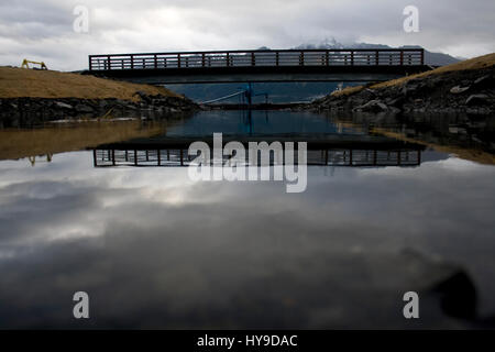 A small simple bridge is reflected in calm water with mountains in background in Seward, Alaska. Stock Photo