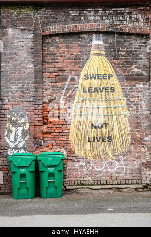A old red brick wall mural painted with a huge broom with words sweep leaves not lives.  Two green garbage cans below it. Stock Photo