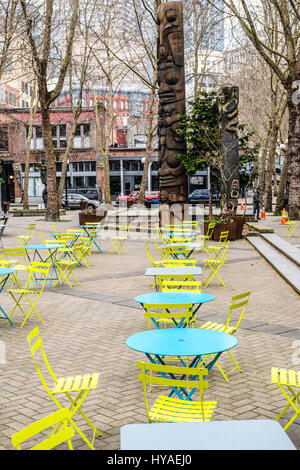 A Northwest Native American totem pole in Seattle's Pioneer Square, along with empty folder tables and chairs for relaxation. Stock Photo