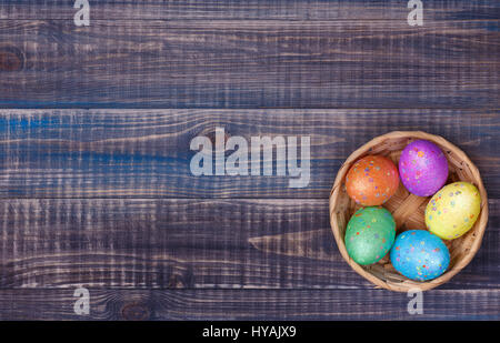 A wicker basket with five colored Easter eggs stands on a wooden background. There is a place for text on the left. Stock Photo