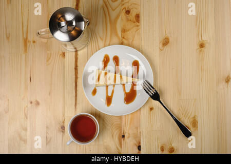 Plate with cheesecake under caramel sauce, a cup of tea and a teapot stand on a light wooden table. Top view Stock Photo