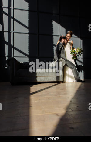 The bride smiling embraces the groom sitting on the arm of the sofa. They are lit by hard light from the window Stock Photo