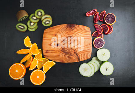 Fresh oranges, kiwi, and apples are sliced and laid out on dark gray surface,  top view with wooden cutting board for text Stock Photo