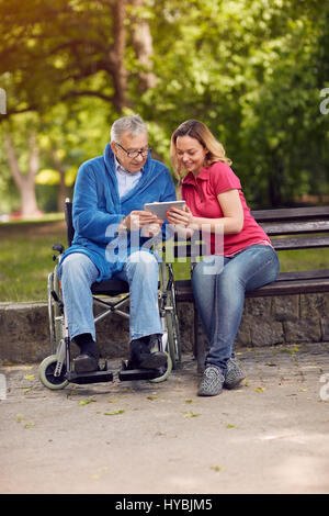 Disabled retiree man in park using tablet with smiling daughter Stock Photo