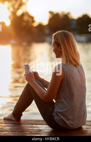 Young female with cellphone relaxing on dock near water Stock Photo