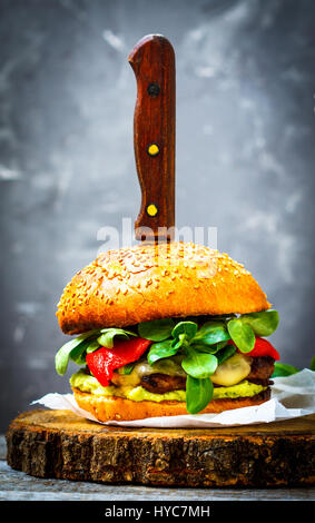 Tasty grilled beef burger with cheese, guacamole and pickled peppers. Dark background, brown paper, wooden stand. Stock Photo