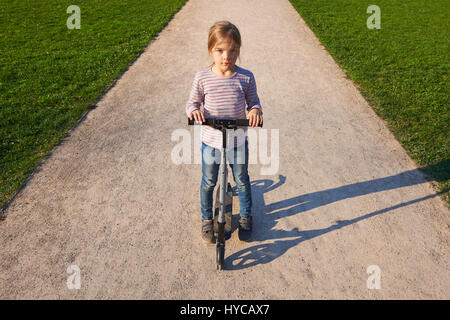 Beautiful child blond girl driving scooter on rural road outdoor in park Stock Photo