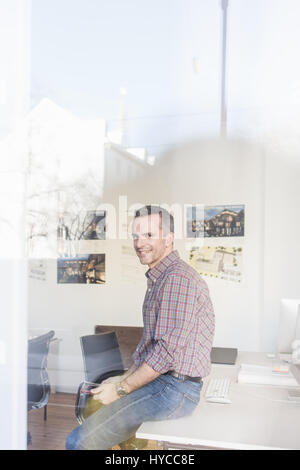 View through glass, side view of mature man sitting on desk looking at camera smiling Stock Photo