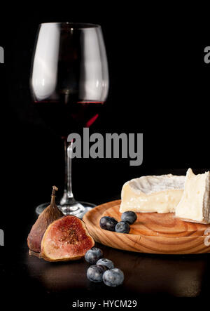 A glass of red wine with fresh figs, blueberries and brie cheese on a wooden board. Black background. Stock Photo