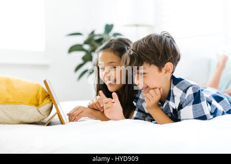 Siblings (10-11, 6-7) lying in bed and using tablet Stock Photo