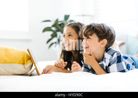 Siblings (10-11, 6-7) lying in bed and using tablet Stock Photo