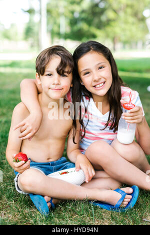 Boy (6-7) and girl (10-11) sitting arm around on grass in summer Stock Photo