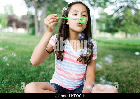 Portrait of girl ( 10-11 ) sitting in meadow and blowing soap bubbles Stock Photo