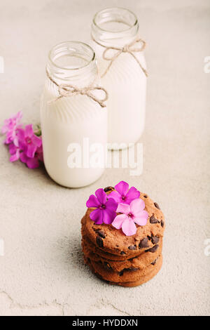 Pile of chocolate chip cookies with two bottles of milk and pink flowers on rustic grey table Stock Photo